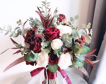 May include: A burgundy and white bridal bouquet with roses, eucalyptus, and other greenery. The bouquet is tied with a burgundy ribbon and white ribbon.