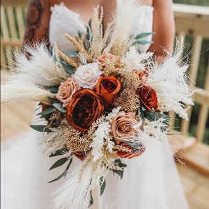 May include: A bridal bouquet featuring a mix of dried flowers and pampas grass. The bouquet includes white, cream, and rust-coloured roses, along with dried grasses and pampas plumes.