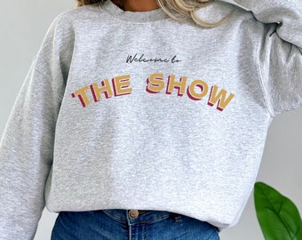 May include: Gray sweatshirt with the text "Welcome to THE SHOW" in yellow and red letters.
