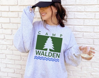 Puede incluir: Una sudadera gris con un pino verde y el texto "CAMP WALDEN" en azul y blanco. La sudadera tiene un diseño de ola azul en la parte inferior.