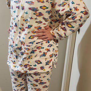 Puede incluir: Un conjunto de pijama blanco de dos piezas con estampado de peces de colores. La parte superior tiene una cremallera y dos bolsillos. Los pantalones son cortos.