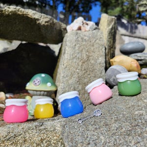 May include: Five small jars with white lids, filled with different colored creams, sitting on a rock surface. The jars are pink, yellow, blue, green, and white. A small clear plastic spoon is in the foreground.