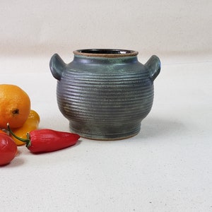 Puede incluir: Una olla de cerámica gris hecha a mano con dos asas. La olla tiene una superficie texturizada y acanalada y un interior oscuro. El borde es de un tono gris más claro. La olla se exhibe con naranjas y pimientos rojos.