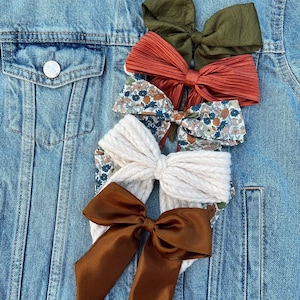 May include: Four different hair bows on a blue denim jacket. The bows are made of different fabrics and colors, including a floral print, a cream knit, a brown satin, and a green and rust colored fabric.