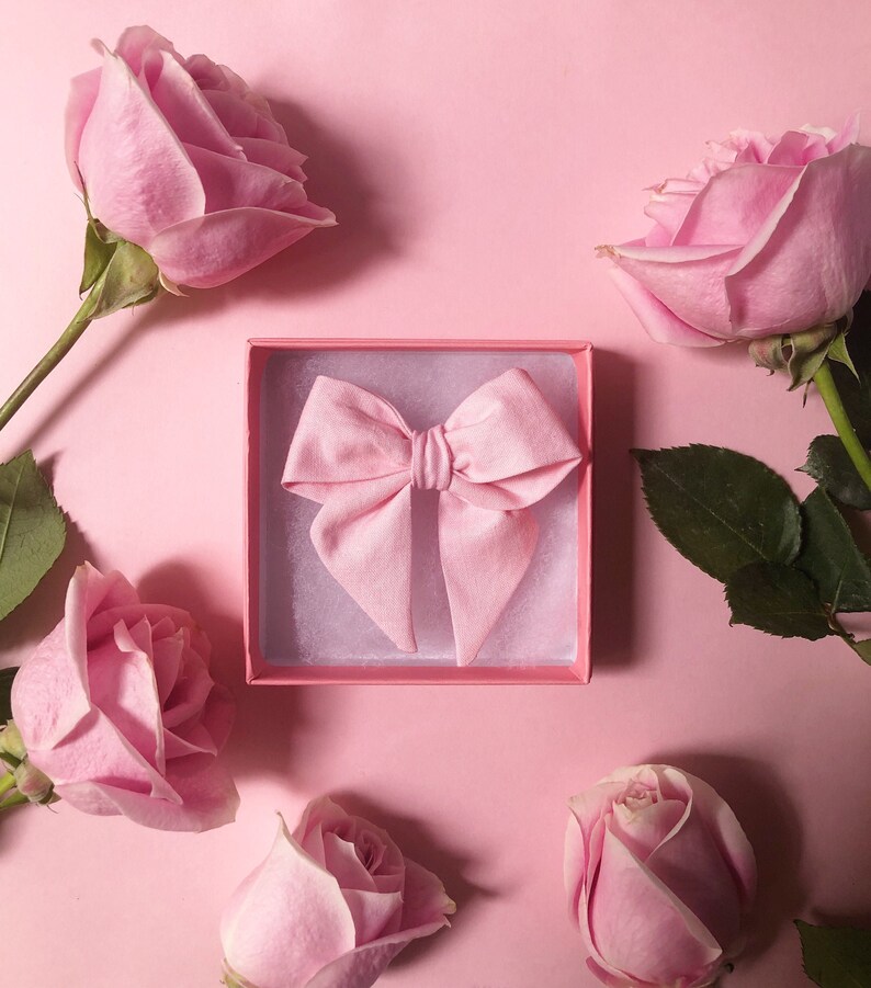 Puede incluir: Un lazo rosa de tela en una caja de regalo rosa, rodeado de rosas rosas sobre un fondo rosa. Las rosas, con sus tallos y hojas verdes, est&aacute;n en diferentes etapas de floraci&oacute;n, creando una est&eacute;tica rom&aacute;ntica.