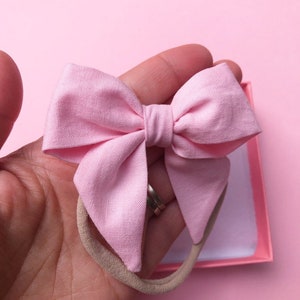 Puede incluir: Un lazo de tela rosa claro sobre una diadema beige. El lazo se sostiene en una mano sobre un fondo rosa. El lazo tiene un dise&ntilde;o sencillo, con un nudo en el centro y dos bucles a cada lado.