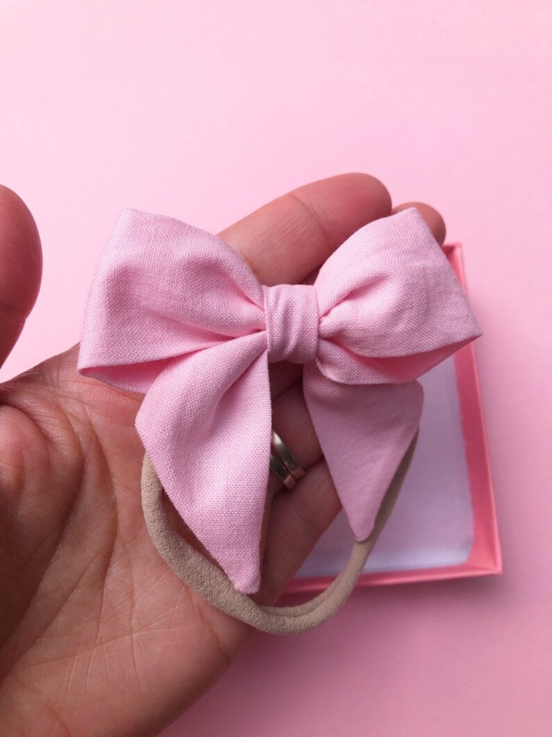 Puede incluir: Un lazo de tela rosa claro sobre una diadema beige. El lazo se sostiene en una mano sobre un fondo rosa. El lazo tiene un dise&ntilde;o sencillo, con un nudo en el centro y dos bucles a cada lado.