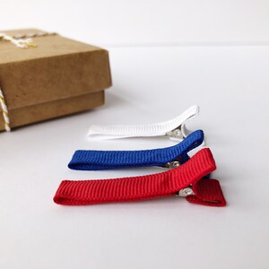 Puede incluir: Tres peque&ntilde;as pinzas para el pelo rojas, blancas y azules, dispuestas sobre una superficie blanca. Las pinzas tienen una textura acanalada y cierres met&aacute;licos. Una caja de regalo marr&oacute;n con una cuerda dorada y blanca est&aacute; en el fondo.