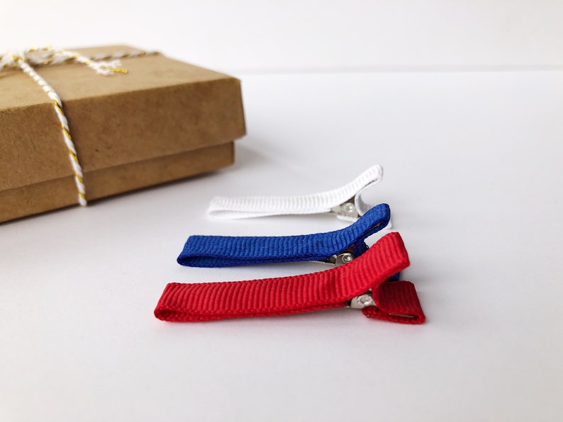 Puede incluir: Tres peque&ntilde;as pinzas para el pelo rojas, blancas y azules, dispuestas sobre una superficie blanca. Las pinzas tienen una textura acanalada y cierres met&aacute;licos. Una caja de regalo marr&oacute;n con una cuerda dorada y blanca est&aacute; en el fondo.