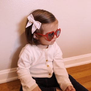 Puede incluir: Un ni&ntilde;o con gafas de sol rojas en forma de coraz&oacute;n y un lazo rosa claro en el pelo. Viste una rebeca blanca con botones dorados y vaqueros azul oscuro. El ni&ntilde;o est&aacute; sentado en un suelo de madera.