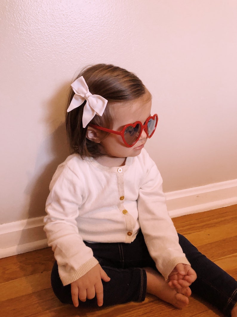 Puede incluir: Un ni&ntilde;o con gafas de sol rojas en forma de coraz&oacute;n y un lazo rosa claro en el pelo. Viste una rebeca blanca con botones dorados y vaqueros azul oscuro. El ni&ntilde;o est&aacute; sentado en un suelo de madera.