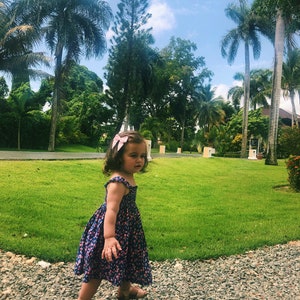 Puede incluir: Una ni&ntilde;a con un vestido azul con estampado floral y sandalias marrones camina por un camino de grava. Un lazo rosa adorna su cabello. El fondo muestra hierba verde, palmeras y un cielo azul brillante.