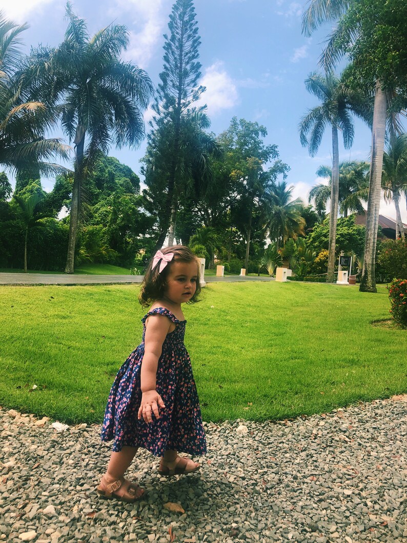 Puede incluir: Una ni&ntilde;a con un vestido azul con estampado floral y sandalias marrones camina por un camino de grava. Un lazo rosa adorna su cabello. El fondo muestra hierba verde, palmeras y un cielo azul brillante.