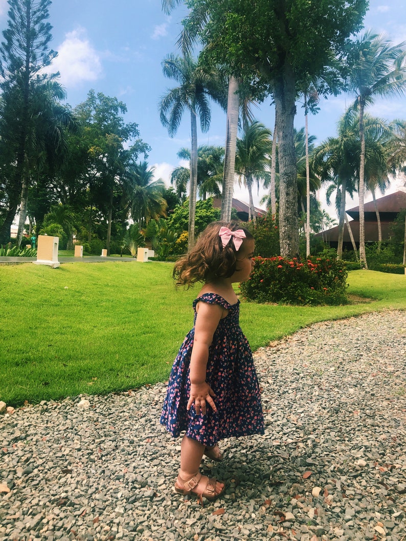 Puede incluir: Una ni&ntilde;a con un vestido azul marino con estampado floral rosa y sandalias marrones, de pie en un camino de grava. Un lazo rosa adorna su cabello. El fondo muestra hierba verde, palmeras y cielo azul. Ambiente soleado.