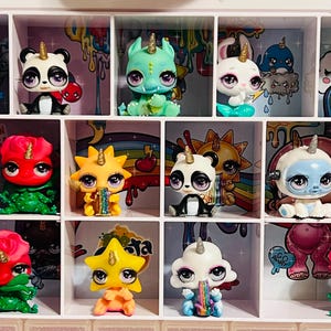 May include: A collection of colorful, unicorn-themed figurines displayed in a pink and white organizer. The figurines feature various animal and plant designs, with glitter accents and large eyes. Each figure has a golden horn.