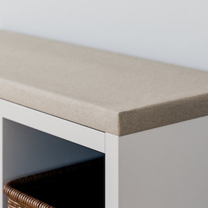 Può includere: Una libreria bianca con un piano superiore in tessuto beige. La parte superiore della libreria è imbottita e sembra una panca. La libreria ha due ripiani e un cesto di vimini sul ripiano inferiore.