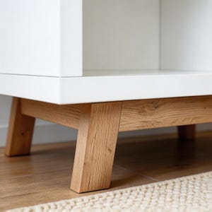Op de afbeelding: Een witte kast met een licht houten onderstel. De kast heeft een eenvoudig, modern design met strakke lijnen. De basis is gemaakt van lichtgekleurd hout en heeft een natuurlijke nerf. De kast staat op een lichtgekleurd tapijt.