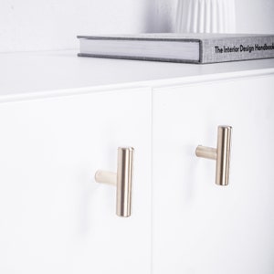 Peut inclure: Deux poignées de tiroir argentées de forme rectangulaire et en forme de T. Les poignées sont montées sur une porte de placard blanche.