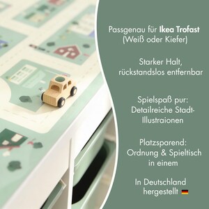 May include: A light green adhesive play mat featuring a detailed city illustration with roads and buildings, applied to a white storage unit. A small wooden toy car sits on the surface. Text includes Passgenau f&uuml;r Ikea Trofast, Starker Halt, r&uuml;ckstandslos entfernbar, Spielspa&szlig; pur, Detailreiche Stadt-Illustrationen, Platzsparend, Ordnung & Spieltisch in einem, In Deutschland hergestellt.