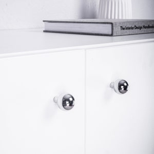 Op de afbeelding: Een witte kast met twee deuren en chromen knopgrepen. De kast staat op een wit oppervlak met een boek erop. Het boek heeft de titel "The Interior Design Handbook".