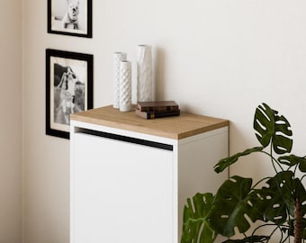 May include: White two-drawer shoe cabinet with a wood top. The cabinet is against a white wall with black and white framed photos and white ceramic vases on top.