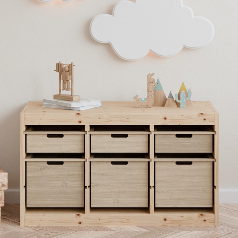 May include: A light-colored wooden storage unit with six drawers and a top shelf. The top shelf displays wooden toys, including a llama, mountains, and a cactus. A cloud-shaped light fixture is mounted on the wall above the unit.