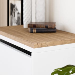 May include: A white cabinet with a wood top and a black trim. The cabinet has a drawer and is sitting on a white surface.