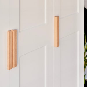Könnte beinhalten: Zwei weiße Schranktüren mit Holzgriffen. Die Griffe sind rechteckig und aus hellem Holz.