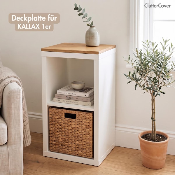 May include: A white square shelving unit with a light wood top panel. The unit features two open compartments, one containing a woven seagrass basket and the other holding three stacked books and a small bowl. Text reads ClutterCover.