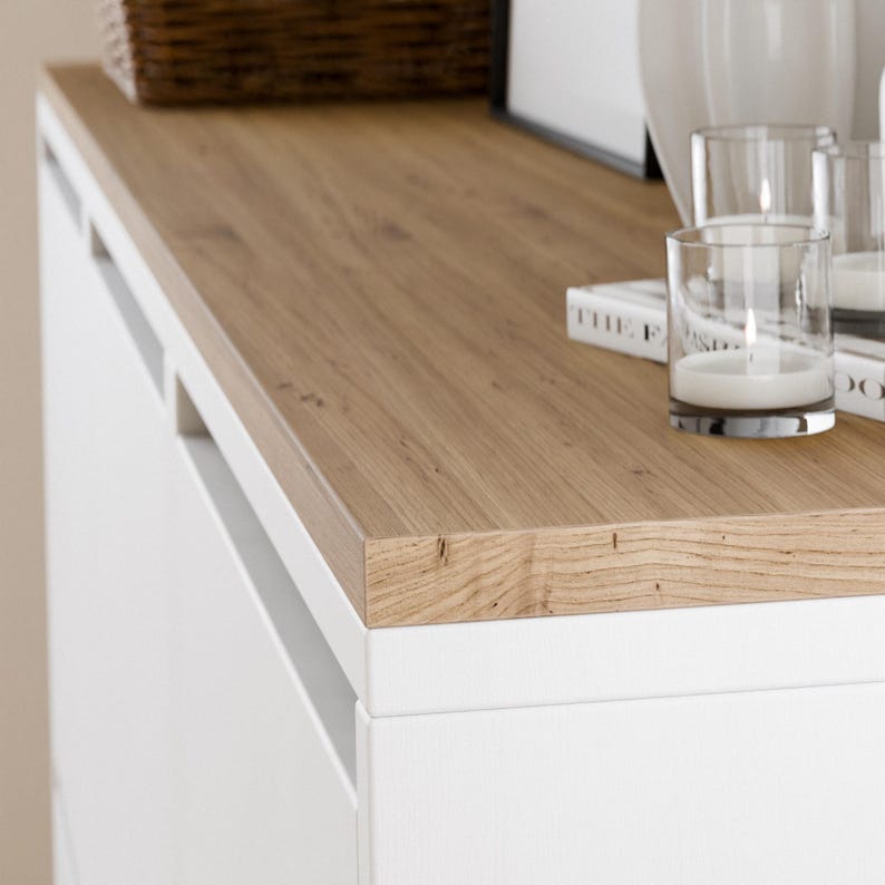 May include: A white cabinet with a wood top. The top is light brown and has a natural wood grain. There are two clear glass candle holders on the top of the cabinet.