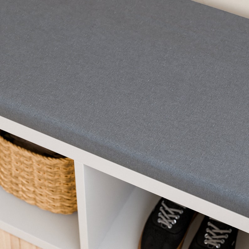 Peut inclure: Un banc de rangement blanc avec un coussin en tissu gris sur le dessus. Le banc a trois &eacute;tag&egrave;res ouvertes, dont l'une contient un panier en osier. Deux paires de chaussures noires sont visibles sous le banc.