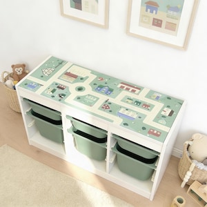 May include: A white rectangular storage unit with three vertical sections, each containing two sage green plastic bins. The top surface features a printed light green play mat design with roads, houses, and trees, providing a flat area for play.