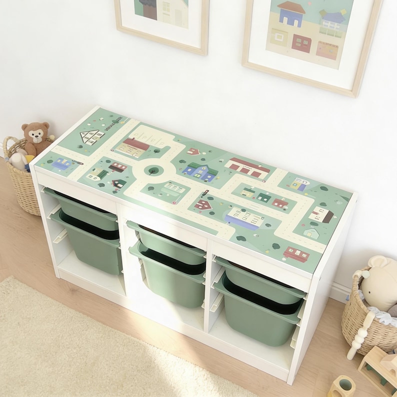 May include: A white rectangular storage unit with three vertical sections, each containing two sage green plastic bins. The top surface features a printed light green play mat design with roads, houses, and trees, providing a flat area for play.