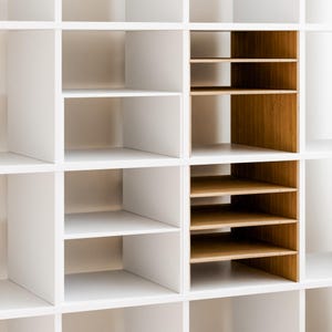 Op de afbeelding: Een witte modulaire opbergkast met meerdere vakken. Sommige planken zijn wit, andere zijn van natuurlijk hout. Het ontwerp heeft een mix van open en gesloten opbergruimtes voor diverse organisatorische behoeften.