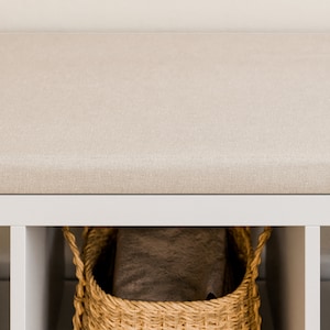 May include: A white bench with a beige fabric cushion and a wicker basket underneath. The basket is filled with a brown blanket.