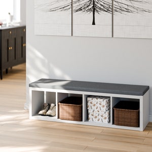 Peut inclure: Banc de rangement blanc avec quatre compartiments. Le banc a un si&egrave;ge rembourr&eacute; gris et est rempli de deux paniers en osier, une paire de chaussures et une bo&icirc;te &agrave; motif de feuilles d'automne.