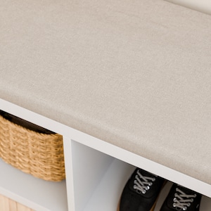 May include: A white storage bench with a beige fabric cushion top. The bench has two open shelves for storage. The shelves are empty.