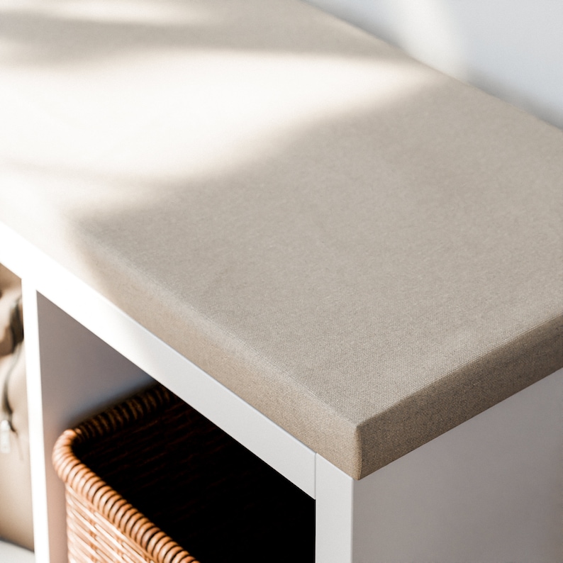 May include: White storage bench with a beige fabric top and a woven basket inside. The bench has a simple design and is perfect for adding storage and seating to any room.