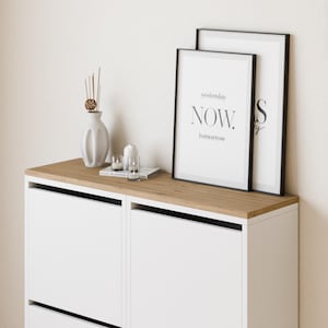 Könnte beinhalten: Ein weißer Schrank mit zwei Schubladen und einer Holzplatte. Zwei schwarze gerahmte Drucke mit dem Text "yesterday NOW. tomorrow" und "S" befinden sich auf der Oberseite des Schranks. Eine weiße Vase mit getrockneten Zweigen und zwei Glaskerzenhalter stehen auf der Oberseite des Schranks.