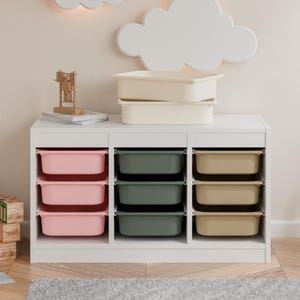 Könnte beinhalten: Ein weißer Aufbewahrungsschrank mit mehreren offenen Fächern, die farbige Kunststoffbehälter enthalten. Die Behälter sind in Rosa, Grün und Beige gehalten. Zwei beige Behälter sind oben auf dem Schrank gestapelt. Ein Holzspielzeug und eine wolkenförmige Leuchte sind sichtbar.