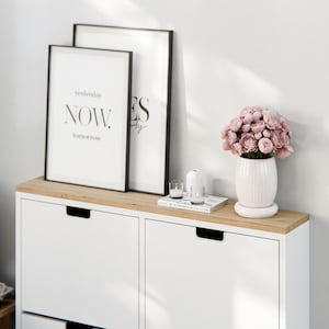 Op de afbeelding: Een witte schoenenkast met twee lades en een houten bovenblad. De kast is versierd met twee zwarte ingelijste prints, een met de tekst "yesterday NOW tomorrow" en de andere met de tekst "YES to" en een bloemstuk in een witte keramische vaas.