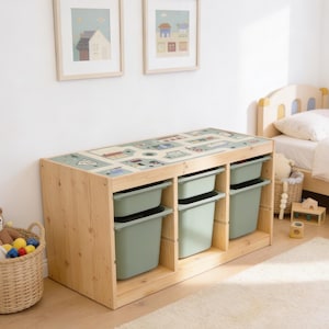 May include: A light-colored wooden storage unit featuring three vertical sections, each containing two sage green plastic bins. The top surface of the unit displays a printed graphic of a town map with roads, buildings, and vehicles in muted tones.