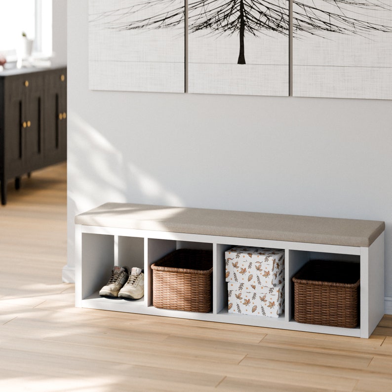 Peut inclure: Banc de rangement blanc avec un dessus rembourr&eacute; et quatre compartiments. Les compartiments contiennent deux paniers en osier, une chaussure et une bo&icirc;te &agrave; motif de feuilles.