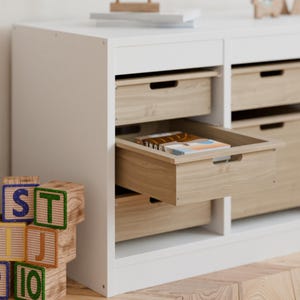 May include: A white storage cabinet with multiple drawers. The drawers are made of light-colored wood and one is partially open, revealing books inside. Wooden alphabet blocks are in the foreground.
