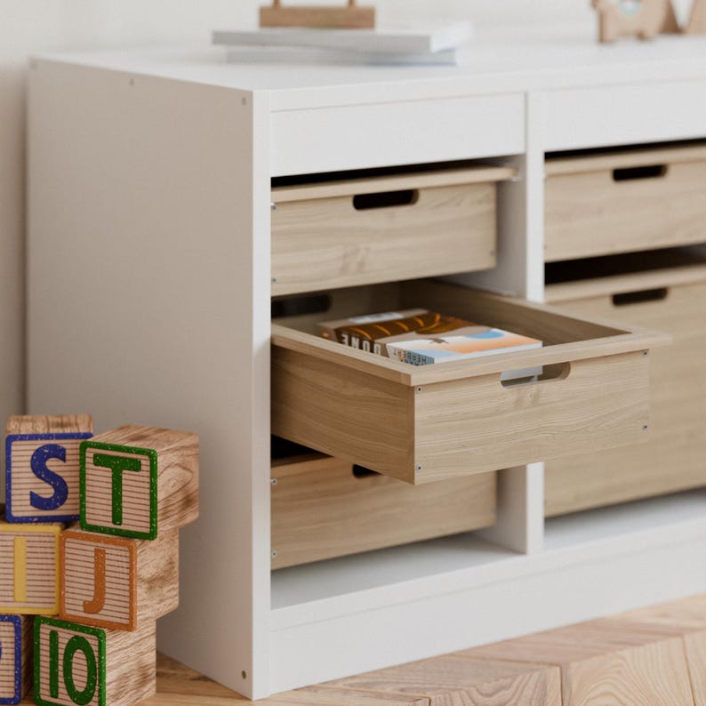May include: A white storage cabinet with multiple drawers. The drawers are made of light-colored wood and one is partially open, revealing books inside. Wooden alphabet blocks are in the foreground.