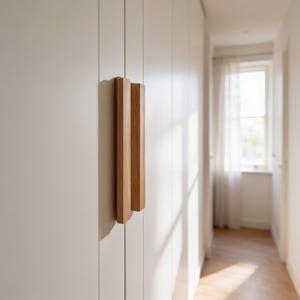 Op de afbeelding: Een verticale rechthoekige houten handgreep met een lichtbruine natuurlijke houtnerfafwerking, bevestigd aan een gladde, matwitte kastdeur. De handgreep heeft een strak, lineair ontwerp met scherpe randen en een vlak oppervlak.