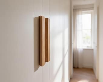 Puede incluir: Un tirador de madera rectangular y vertical con un acabado de veta de madera natural en color marrón claro, fijado a una puerta de armario blanca mate y lisa. El tirador presenta un diseño lineal limpio con bordes afilados y superficie plana.