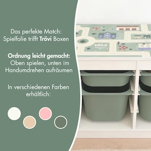 May include: A white storage unit with sage green bins and a top surface featuring a printed town map. Text on the left reads: Das perfekte Match: Spielfolie trifft Tr&ouml;vi Boxen, Ordnung leicht gemacht: Oben spielen, unten im Handumdrehen aufr&auml;umen, In verschiedenen Farben erh&auml;ltlich, with four color circles below.