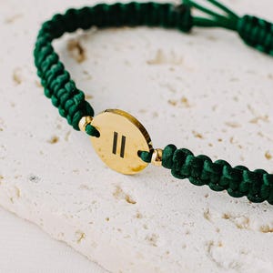 May include: A green macrame bracelet with a gold-colored oval charm. The charm features two vertical lines. The bracelet is adjustable with a sliding knot closure. The bracelet is displayed on a textured, off-white surface.