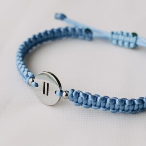 May include: A light blue macrame bracelet with a silver-colored circular charm. The charm features two vertical lines. The bracelet is adjustable and has a sliding knot closure. The bracelet is a light blue color.